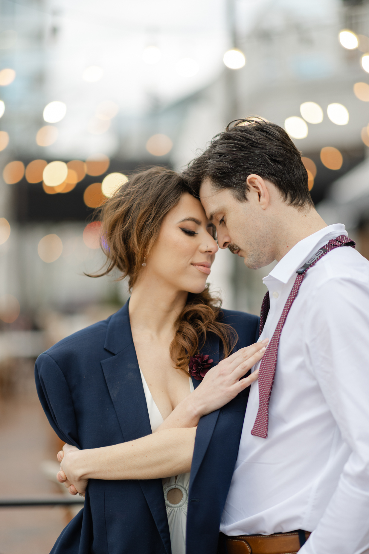Engagement photography of a couple standing on the street in Virginia Beach downtown in front of the outdoor seating of the restaurant with their foreheads touching. The bride is wearing  a white dress and a groom's jacket 