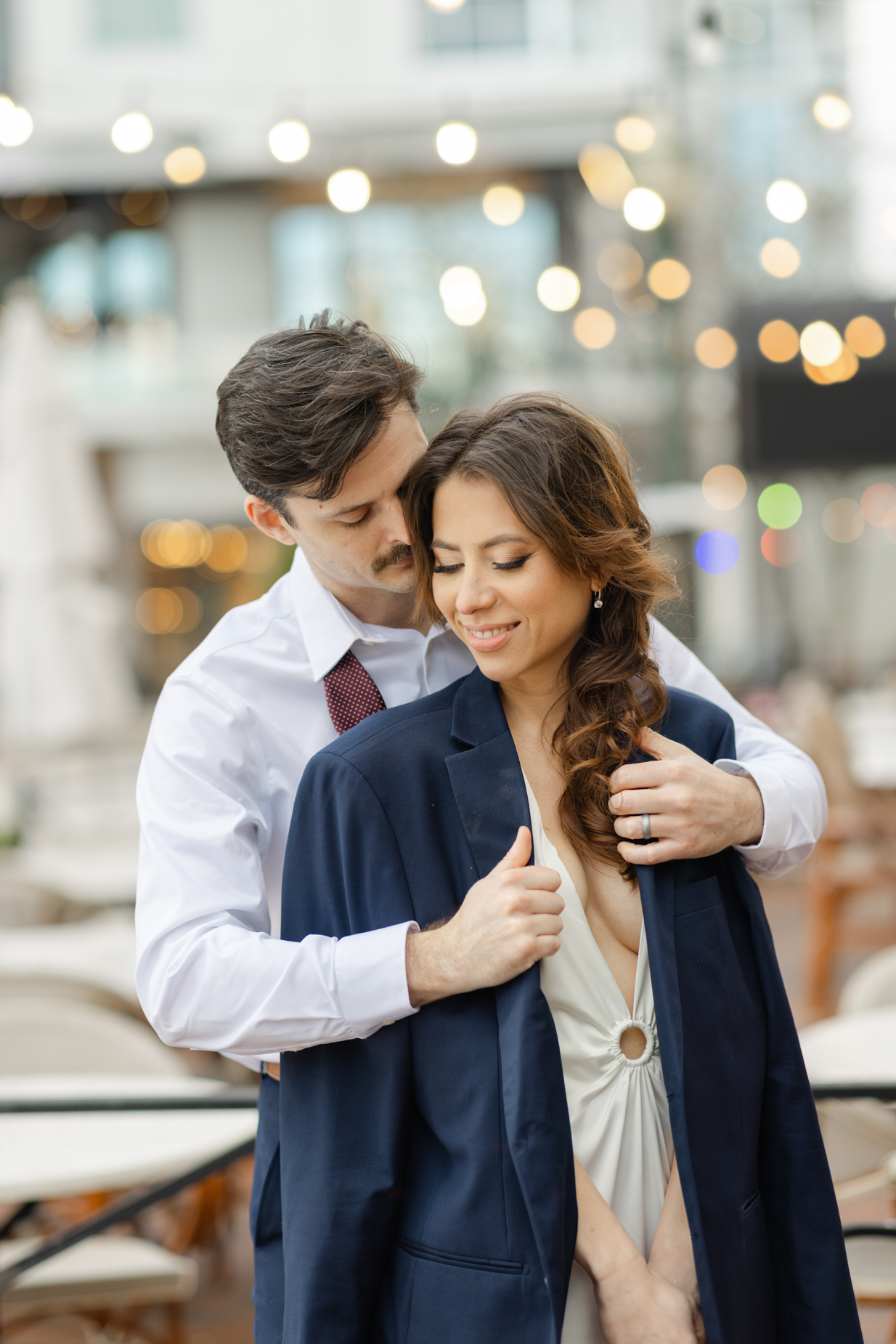 Engagement photography of a couple standing on the street in Virginia Beach downtown in front of the outdoor seating of the restaurant. The groom throws a jacket over the bride's shoulders 