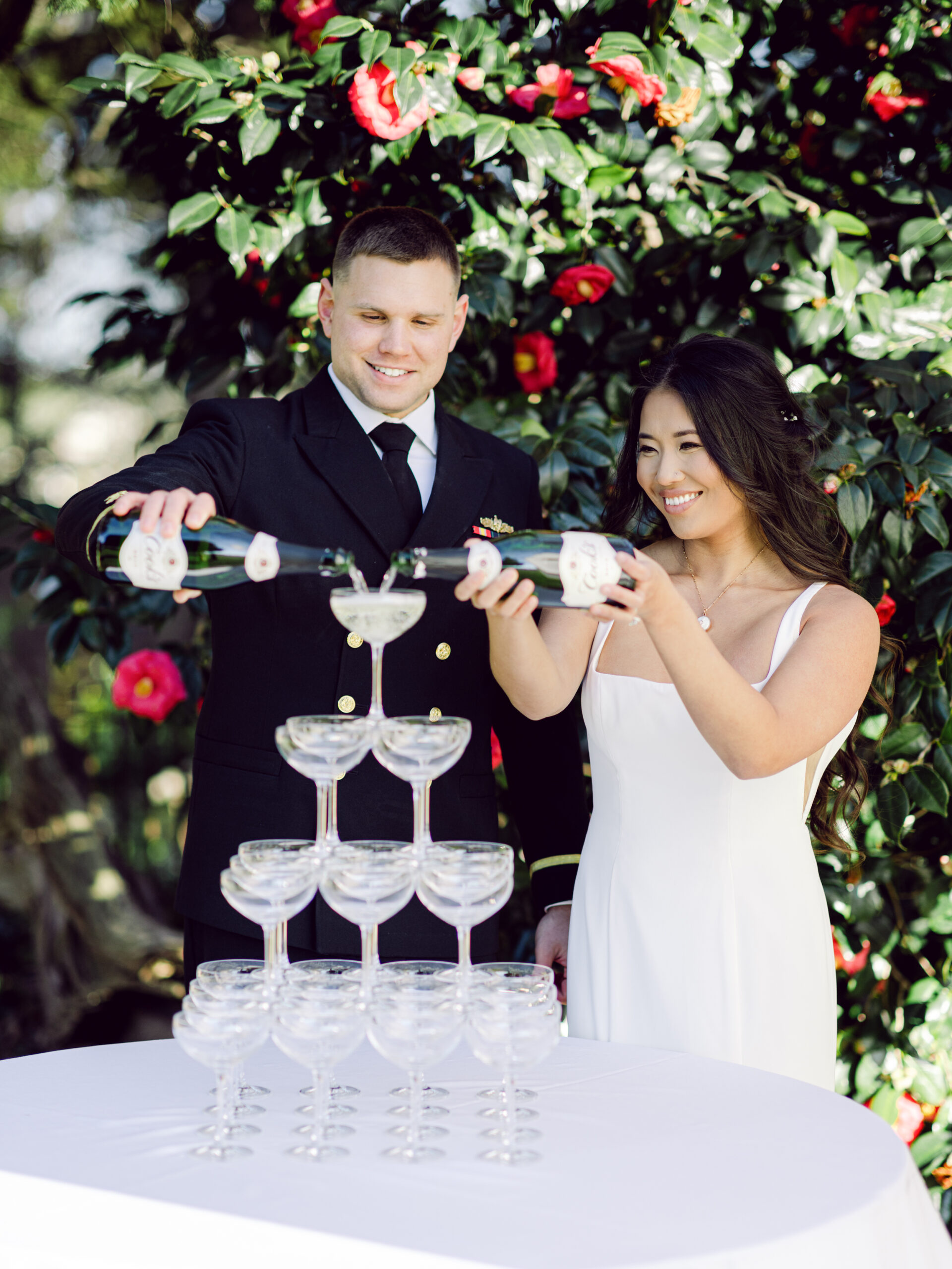 Hermitage Museum and Gardens  wedding photography of a bride and groom standing in front of a bush with red flowers and pouring champagne into champagne tower taken by Virginia Wedding photographers Glenn and Nadya